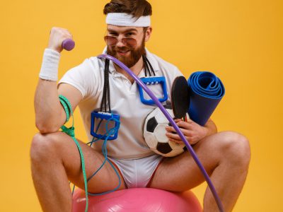 Portrait of a satisfied sports man sitting on a fitness ball and doing exercises with dumbbell isolated on a orange background